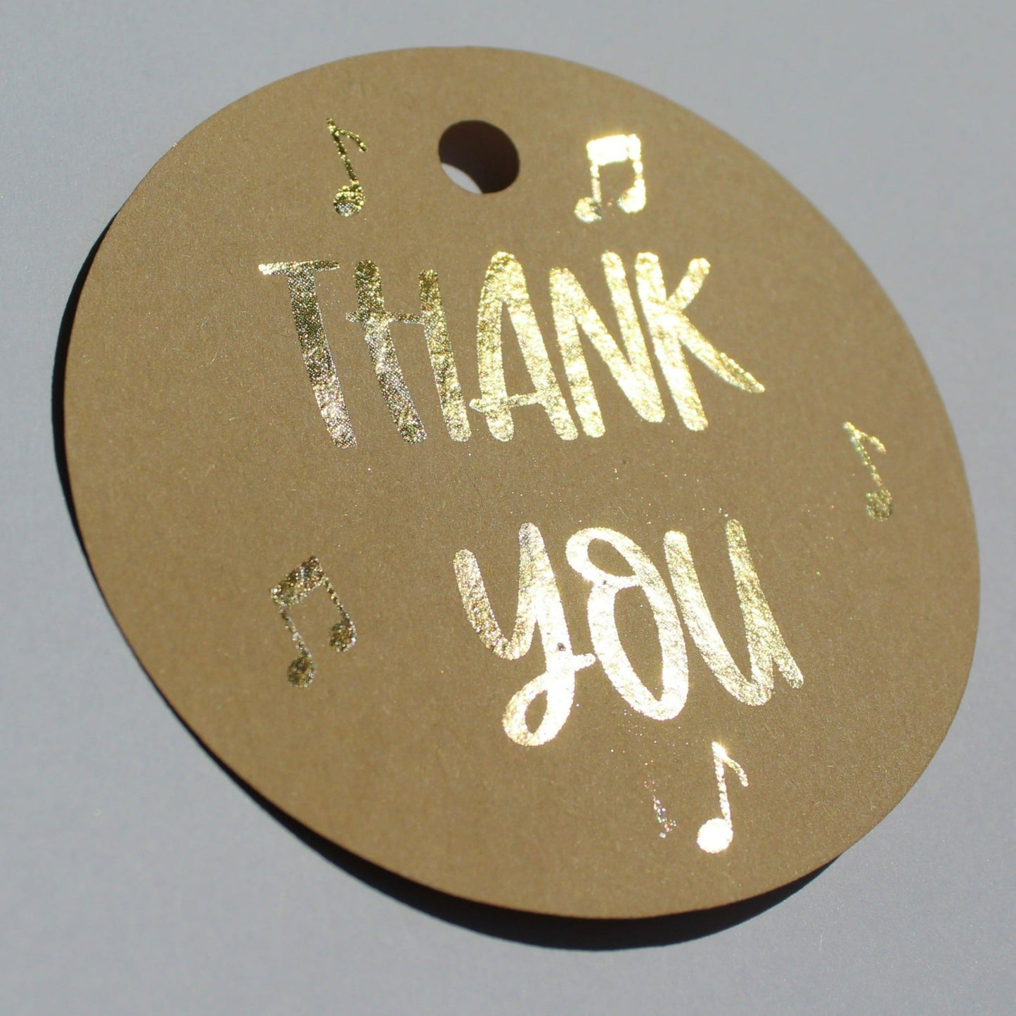 Round brown kraft gift tag. Text reads 'thank you' in gold shiny foil with several music notes around the text.