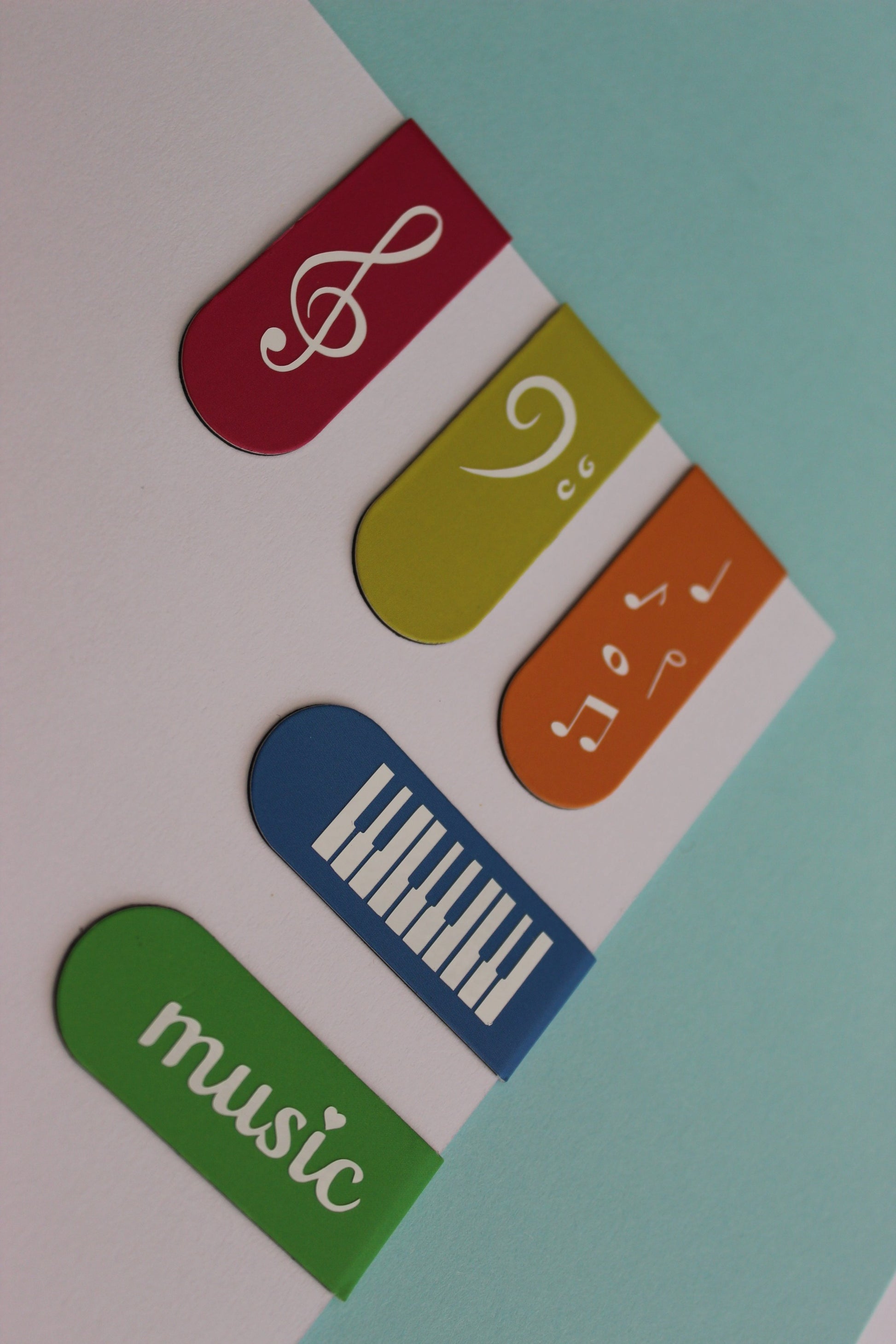 5 bookmarks on a piece of card. Pink, blue, yellow, green and orange. Each bookmark has a different symbol; treble clef, piano keys, bass clef, 'music' and music notes.