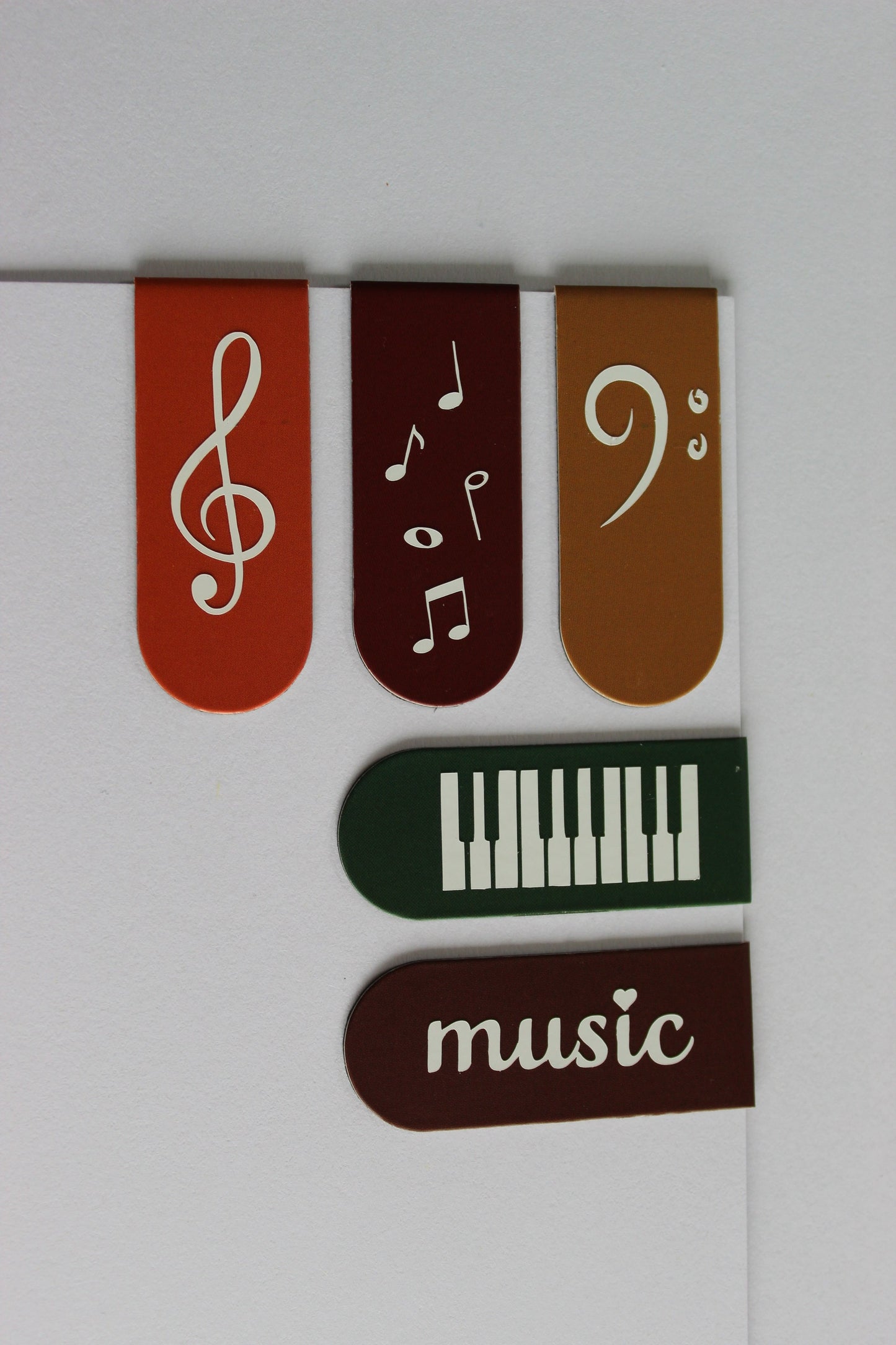 5 bookmarks on a piece of card. Orange, light brown, mustard, green and dark brown. Each bookmark has a different symbol; treble clef, piano keys, bass clef, 'music' and music notes.