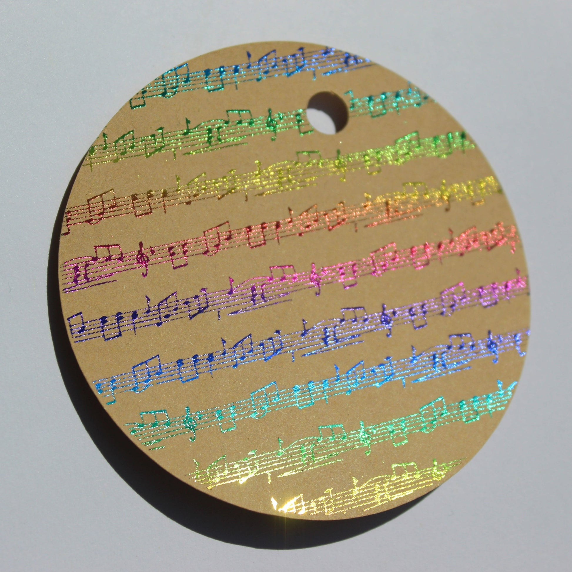 Round brown kraft gift tag. Design of rows of music in a multicoloured shiny foil.