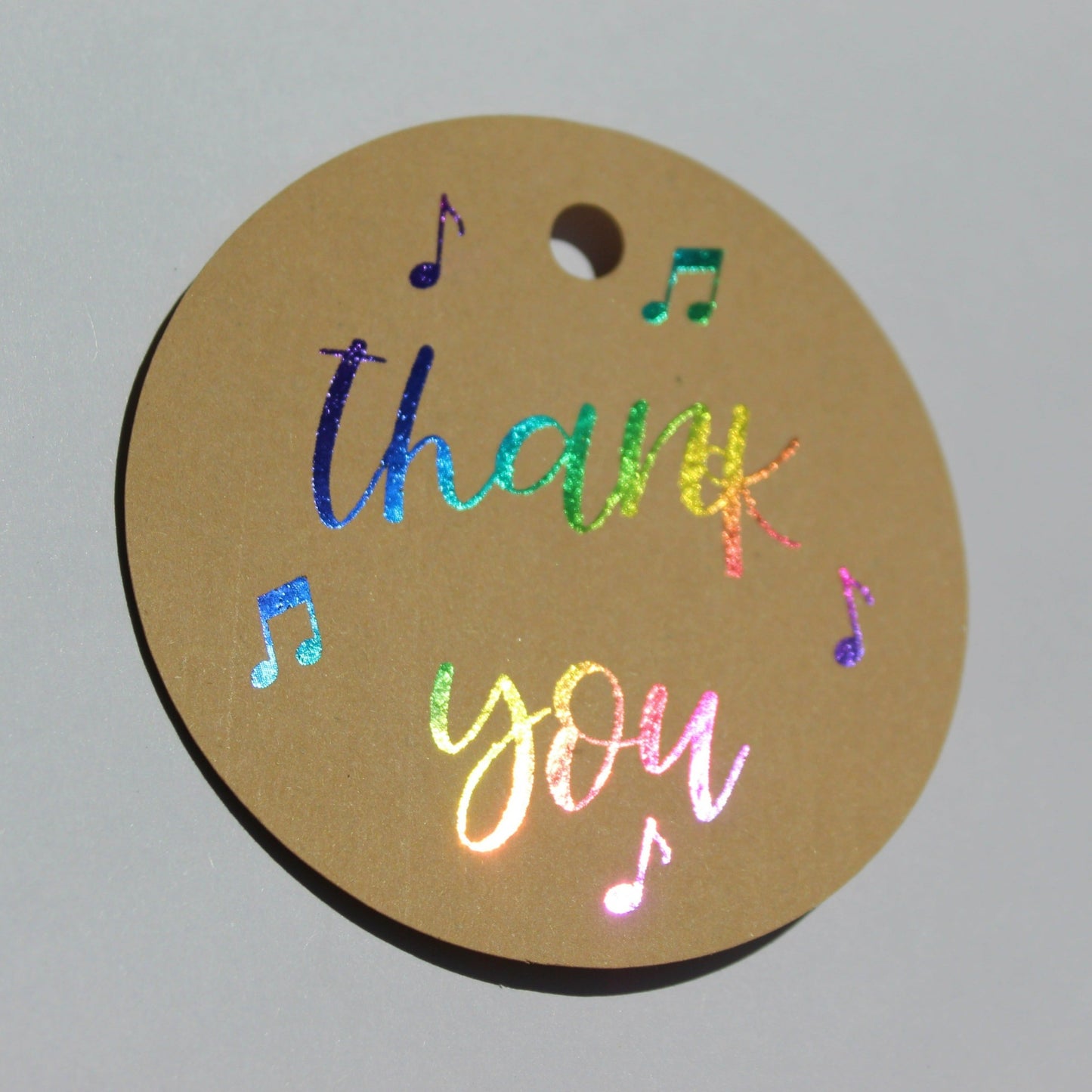Round brown kraft gift tag. Text reads 'thank you' in multicoloured shiny foil with several music notes around the text.