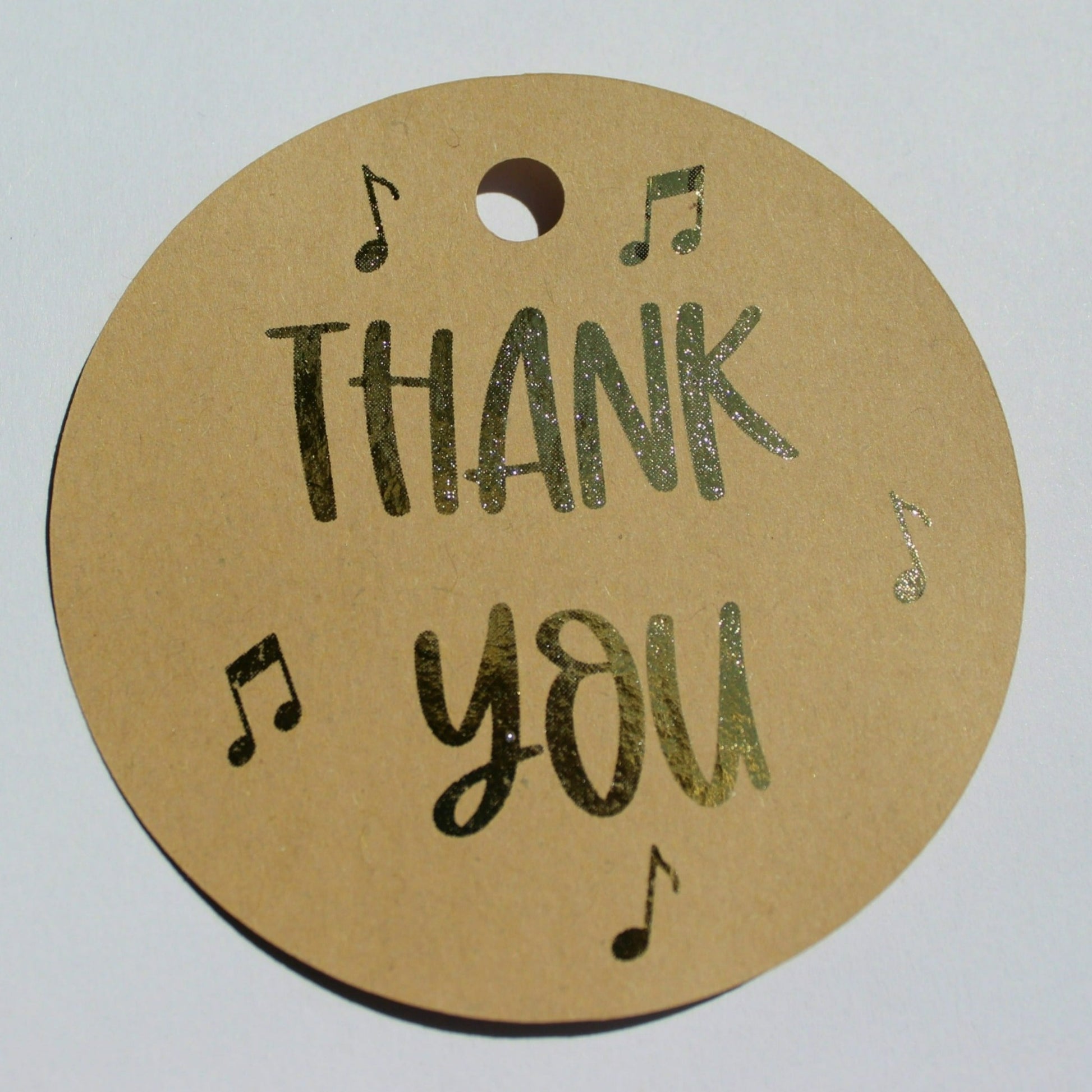 Round brown kraft gift tag. Text reads 'thank you' in gold shiny foil with several music notes around the text.