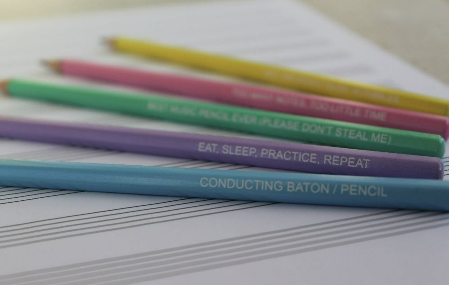 Fading out picture showing pastel coloured pencils on some manuscript paper. Pencil slogans read; Conducting baton/pencil, eat sleep practice repeat, best music pencil ever (please don't steal me), too many notes too little time, very important music scribbles.