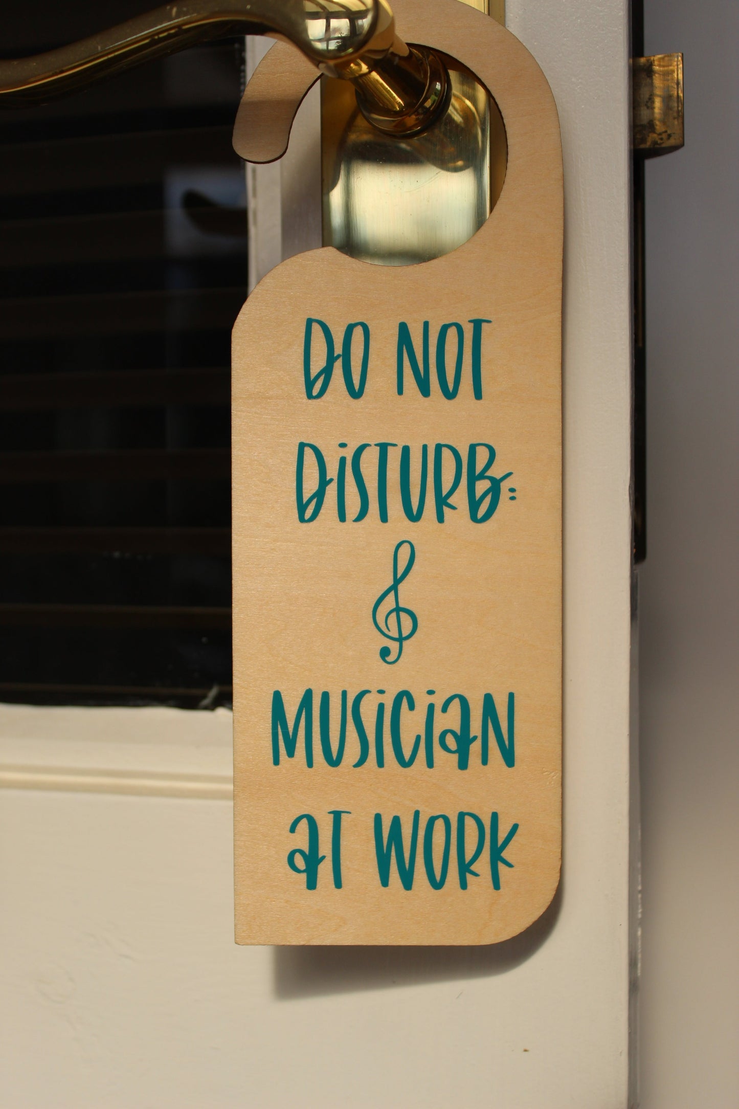 A wooden door hanger on a door handle. Displays the text ' Do not disturb: musician at work' in teal vinyl