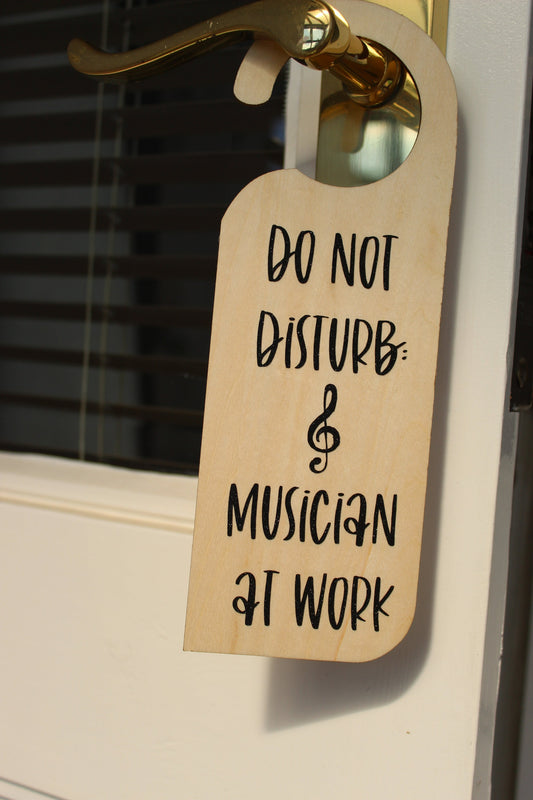 A wooden door hanger on a door handle. Displays the text ' Do not disturb: musician at work' in black shimmery vinyl