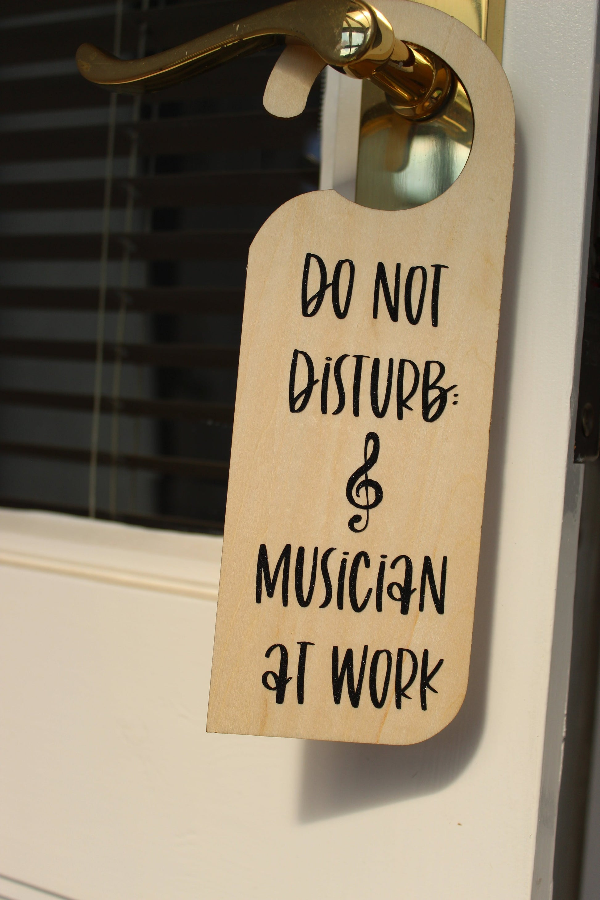 A wooden door hanger on a door handle. Displays the text ' Do not disturb: musician at work' in black shimmery vinyl