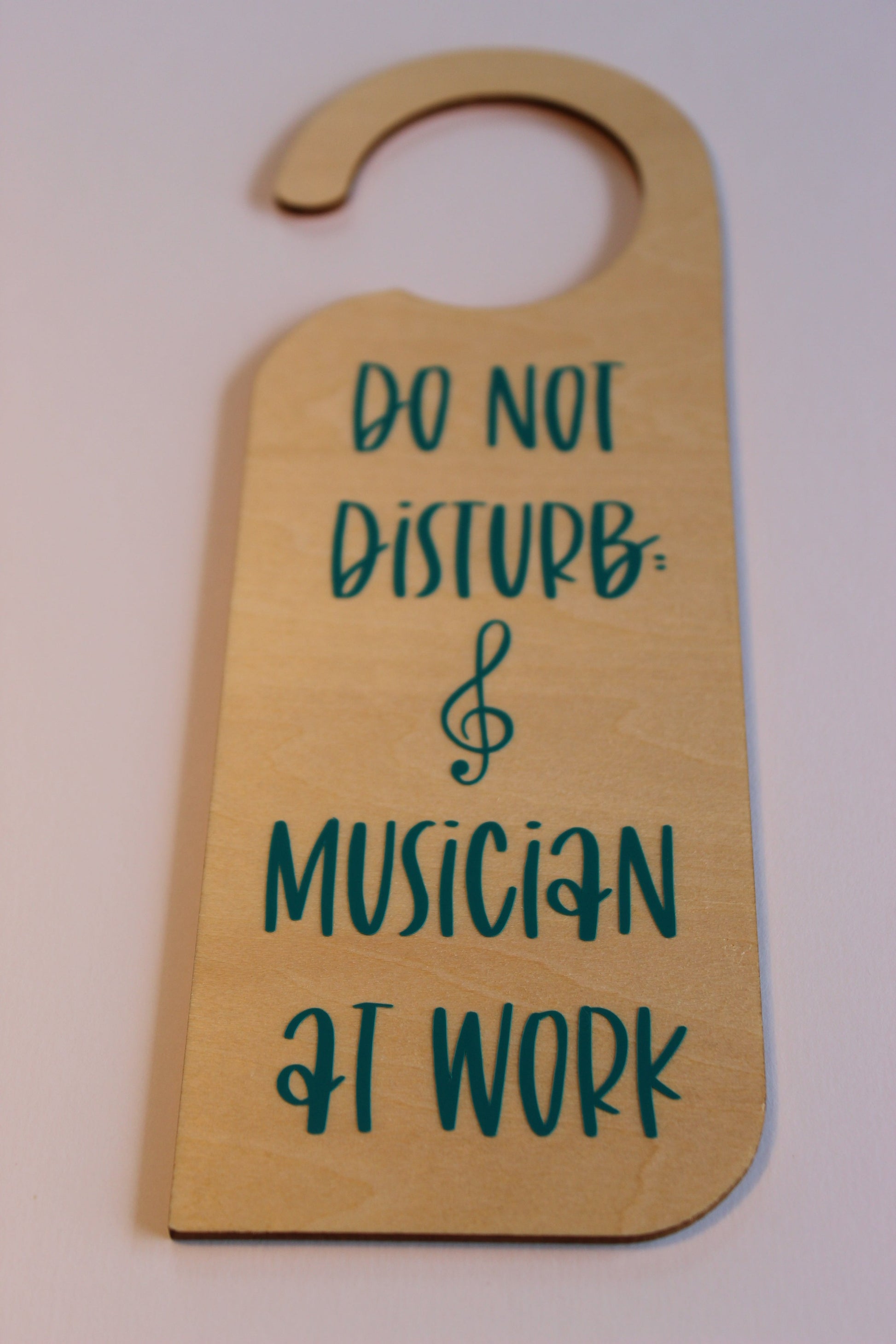 A wooden door hanger, displaying the text 'Do not disturb: musician at work' in teal vinyl