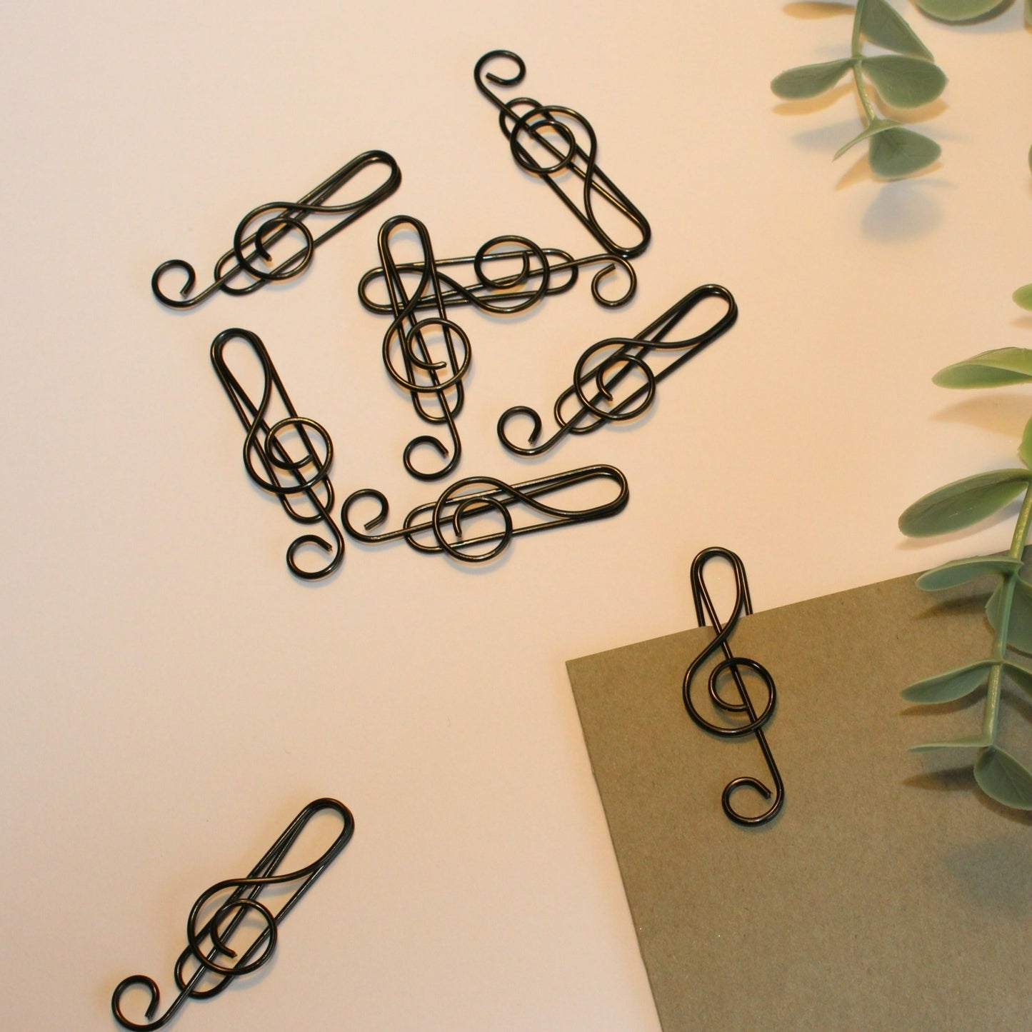 Several black treble clef paperclips. Green foliage and grey card on a white background.