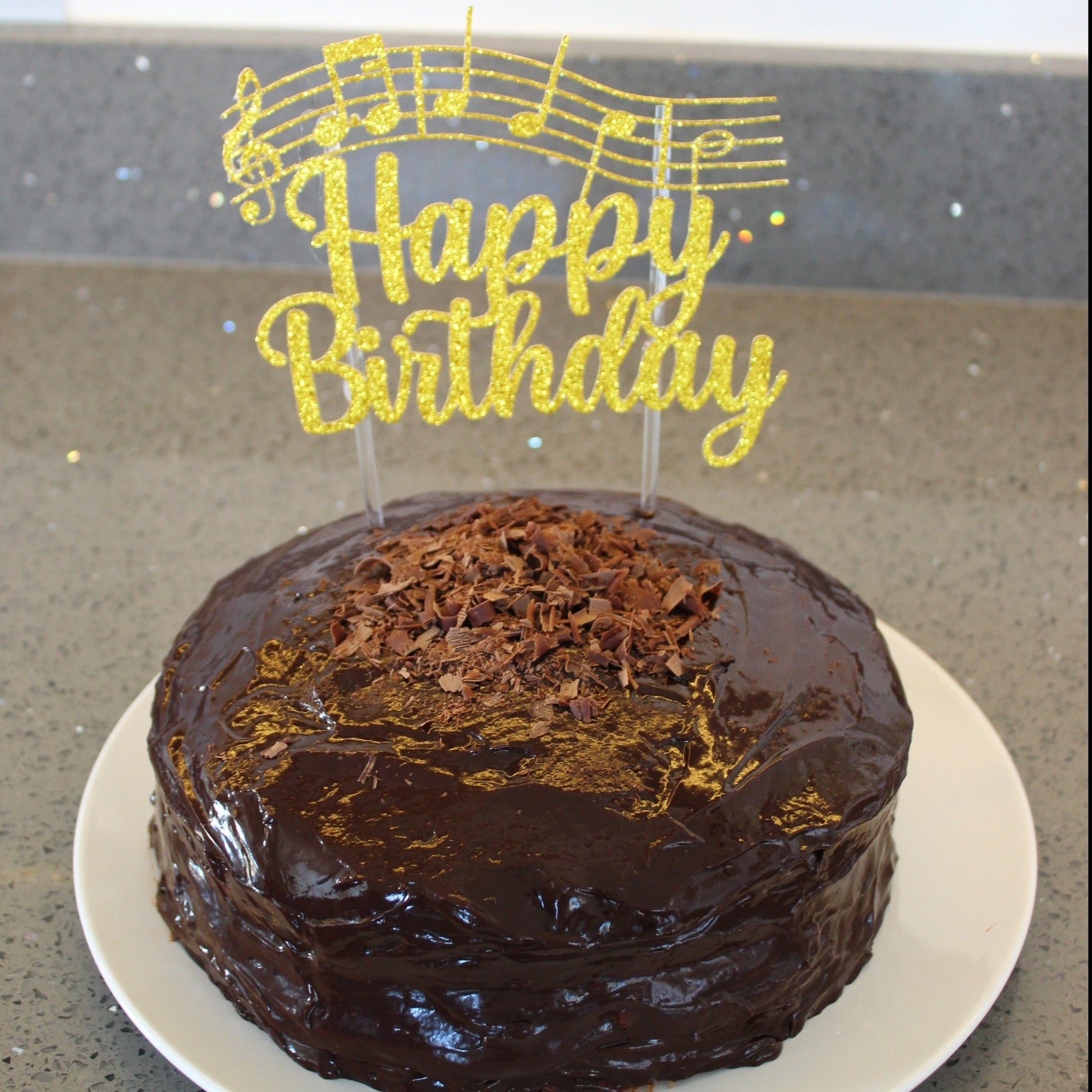 Picture of a cake with a gold glittery cake topper. Cake topper reads 'happy birthday' and shows the notes of the song 'happy birthday to you'