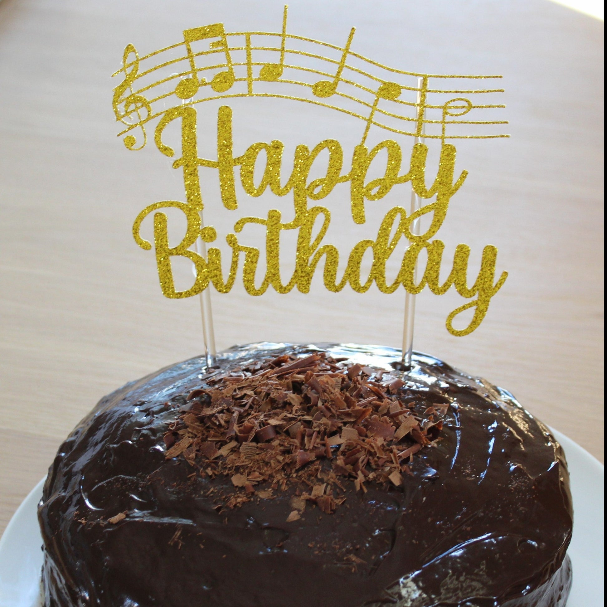 Picture of a cake with a gold glittery cake topper. Cake topper reads 'happy birthday' and shows the notes of the song 'happy birthday to you'